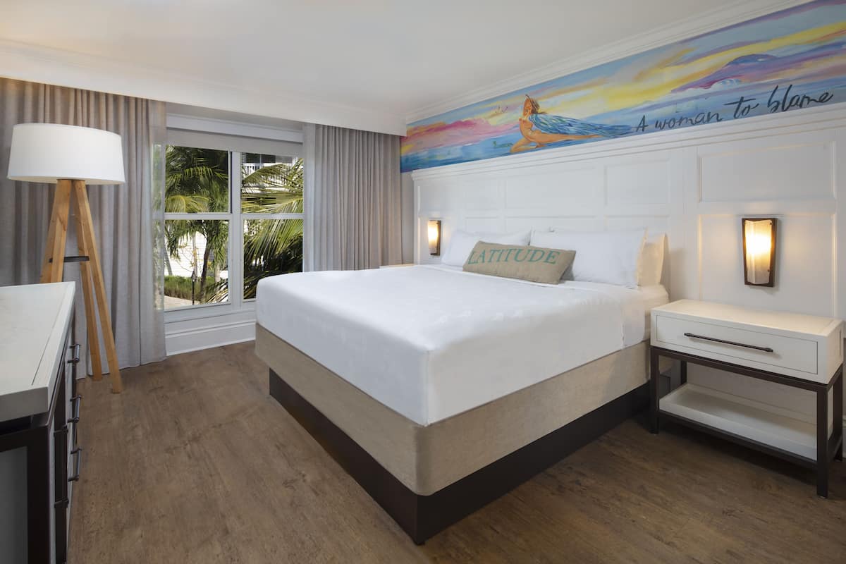A hotel room with a large bed, a bedside table with a lamp, a window with curtains, and a mural on the wall.