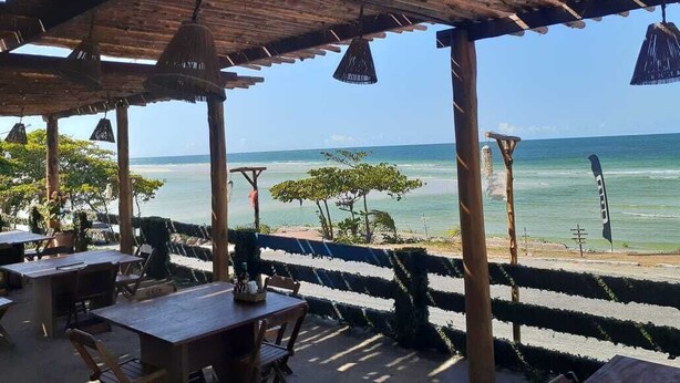 An outdoor seating area with wooden tables and chairs, a pergola with hanging lamps, and a view of the ocean. - Hotel Salinopolis e Kitepoint (Salinópolis)