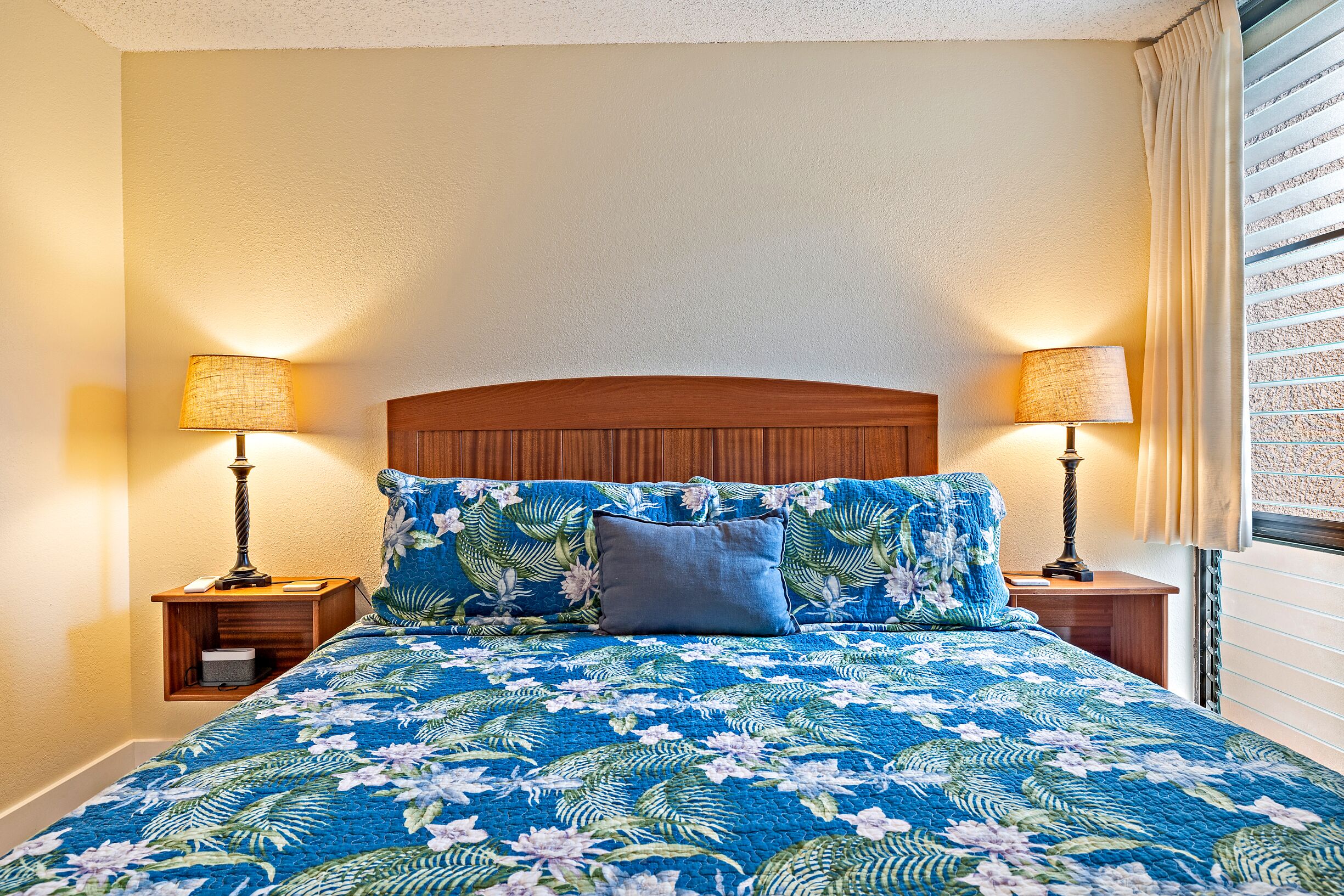 A bed with a floral bedspread, two nightstands with lamps, and a window with blinds.