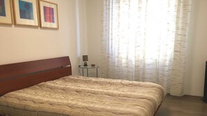 Ett sovrum med en säng i trä, ett litet sängbord, en lampa och ett fönster med tunna, genomskinliga gardiner.