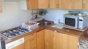 A compact kitchen with wooden cabinets, a gas stove, a microwave, a sink, and a small shelf with kitchen items.