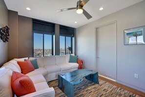A modern living room with a white sofa, colorful cushions, a wooden coffee table, and large windows offering a city view.