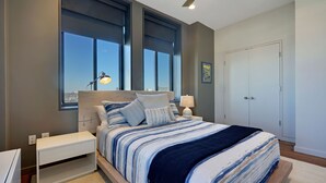 Een moderne slaapkamer met een groot bed, nachtkastjes, een raam met jaloezieën en een deur.