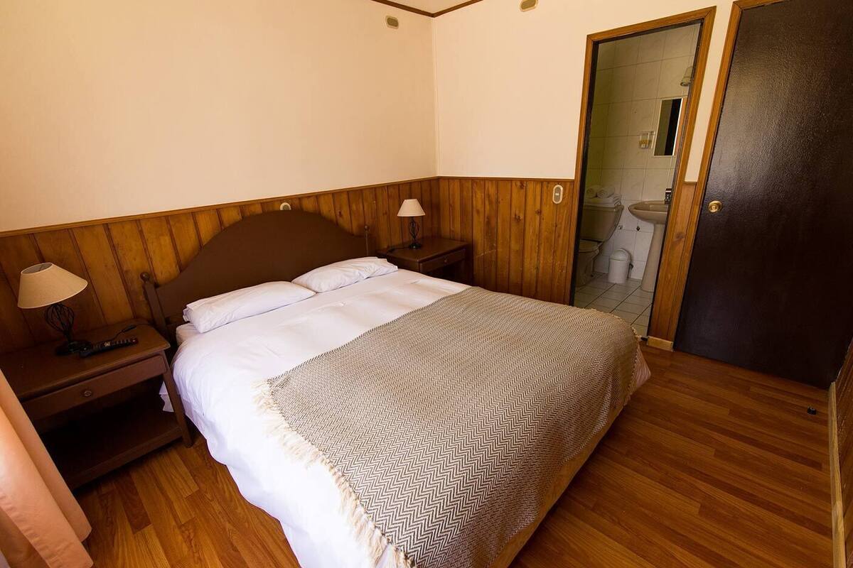 A bedroom with a wooden headboard, a bed with white linens, a patterned throw blanket, a nightstand with a lamp, and a door leading to a bathroom.