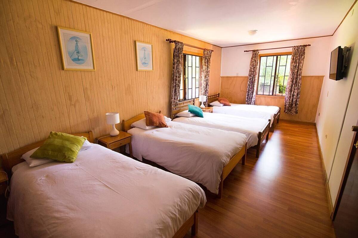 A hotel room with three single beds, wooden flooring, and a large window with curtains.