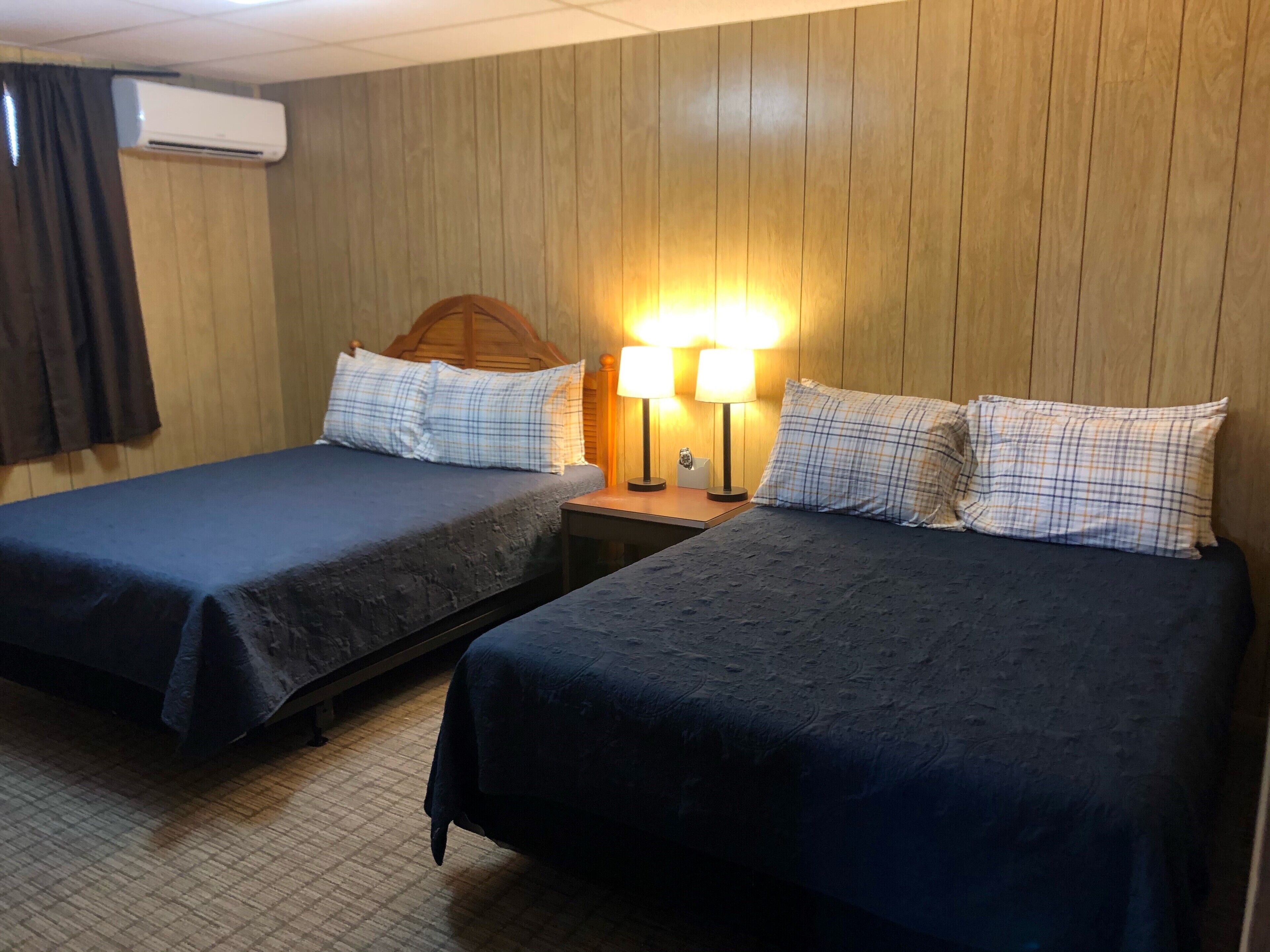 Une chambre d’hôtel avec deux lits, des murs lambrissés et une table de chevet avec des lampes, ainsi qu’un appareil de climatisation.