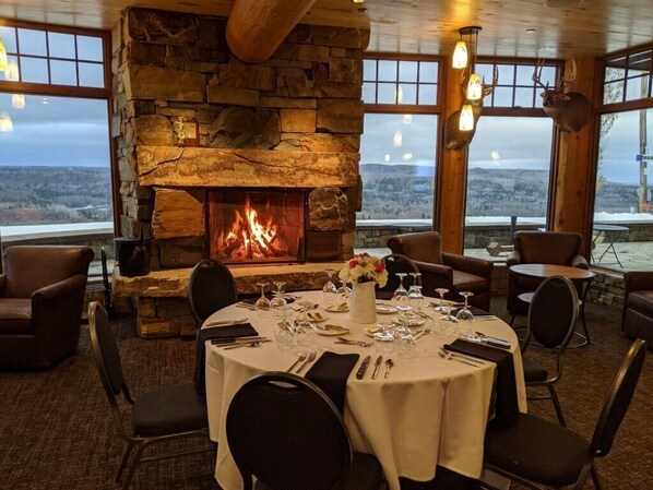 A dining area with a stone fireplace, round tables set for four, and a view of the landscape through large windows.