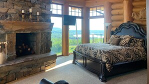 A bedroom with a stone fireplace, a large bed with patterned bedding, and a view of the outdoors. - Mont du Lac Resort (Superior)