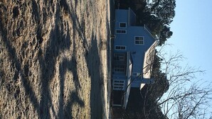 Een twee verdiepingen tellend huis met een puntige dak en een veranda, met aan de rechterkant een grote boom.