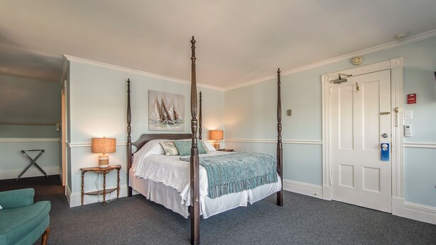 A four-poster bed with a white bedspread and blue accents, a wooden nightstand with a lamp, a teal armchair, and a door with a fire alarm.