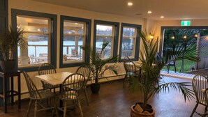 A cozy indoor seating area with wooden tables and chairs, large windows overlooking a snowy outdoor scene, and potted plants adding greenery. - The Tuna Blue Inn and Restaurant (Hubbards)