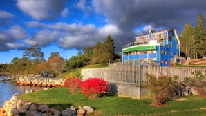A multi-story house with a blue exterior, located near a body of water, surrounded by a wooden fence, and featuring a stone wall and a grassy area with red foliage. - The Tuna Blue Inn and Restaurant (Hubbards)