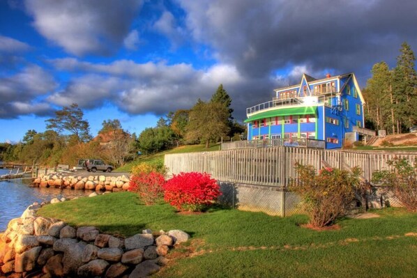 A multi-story house with a blue exterior, located near a body of water, surrounded by a wooden fence, and featuring a stone wall and a grassy area with red foliage.