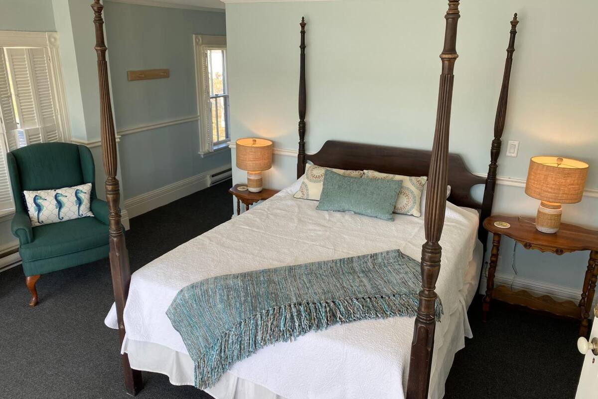 A four-poster bed with a white bedspread and a teal throw blanket, flanked by two wooden nightstands with lamps, in a room with light blue walls and a green armchair.