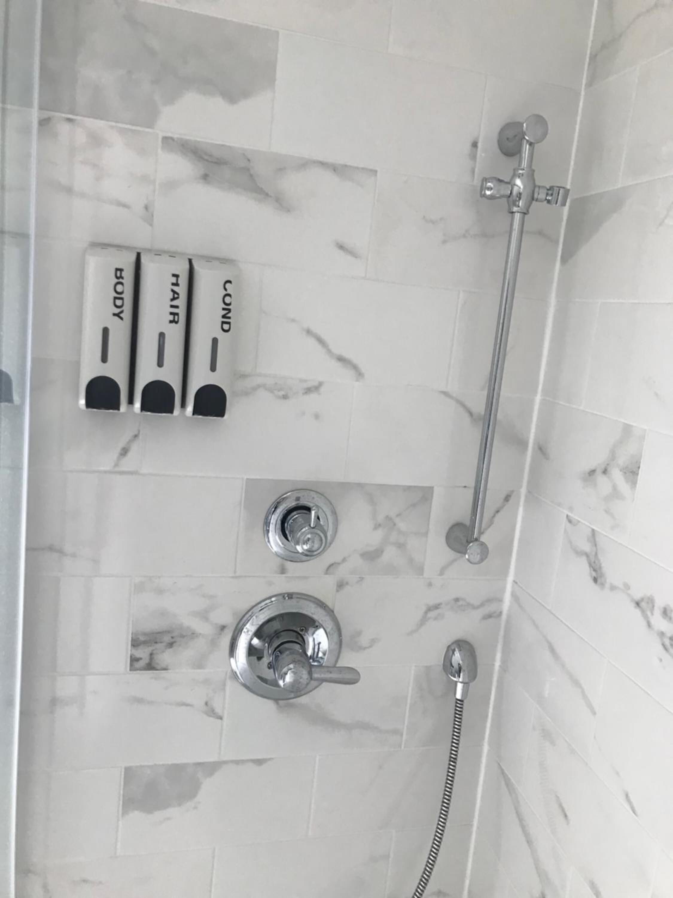 Ein Badezimmer mit Dusche, Duschbrause und zwei Seifenspendern, beschriftet mit „KÖRPER“ und „HAAR“.