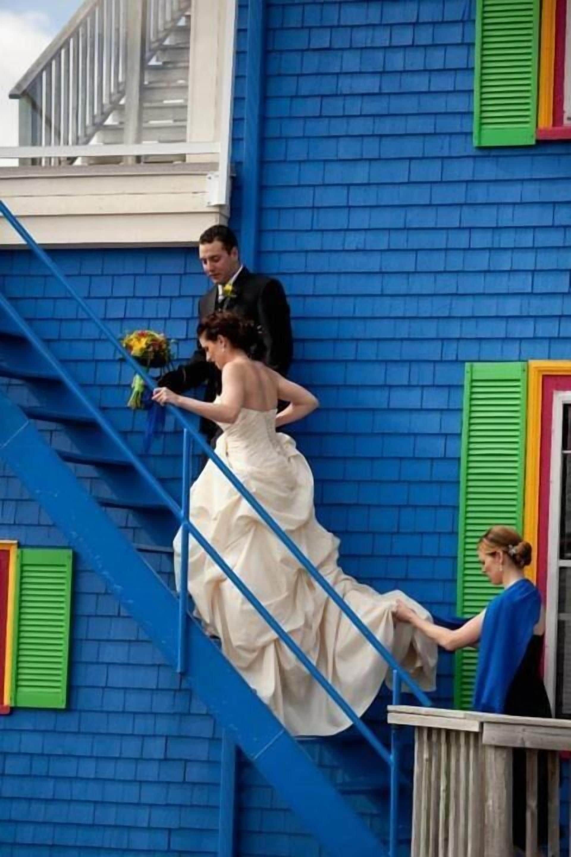A bride in a white dress climbing a blue staircase with a man in a suit and a woman assisting her.
