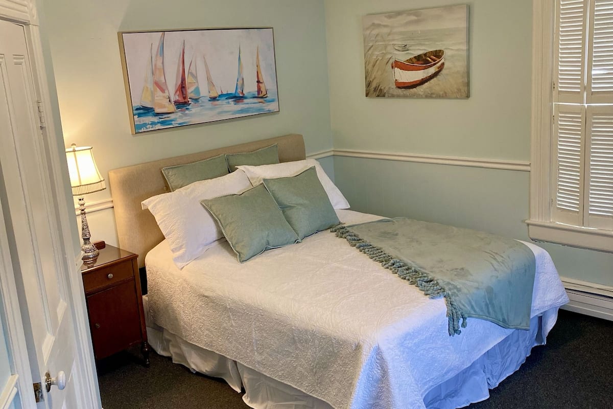 A bedroom with a bed, a nightstand, a lamp, a painting of sailboats, and a painting of a boat on the wall.