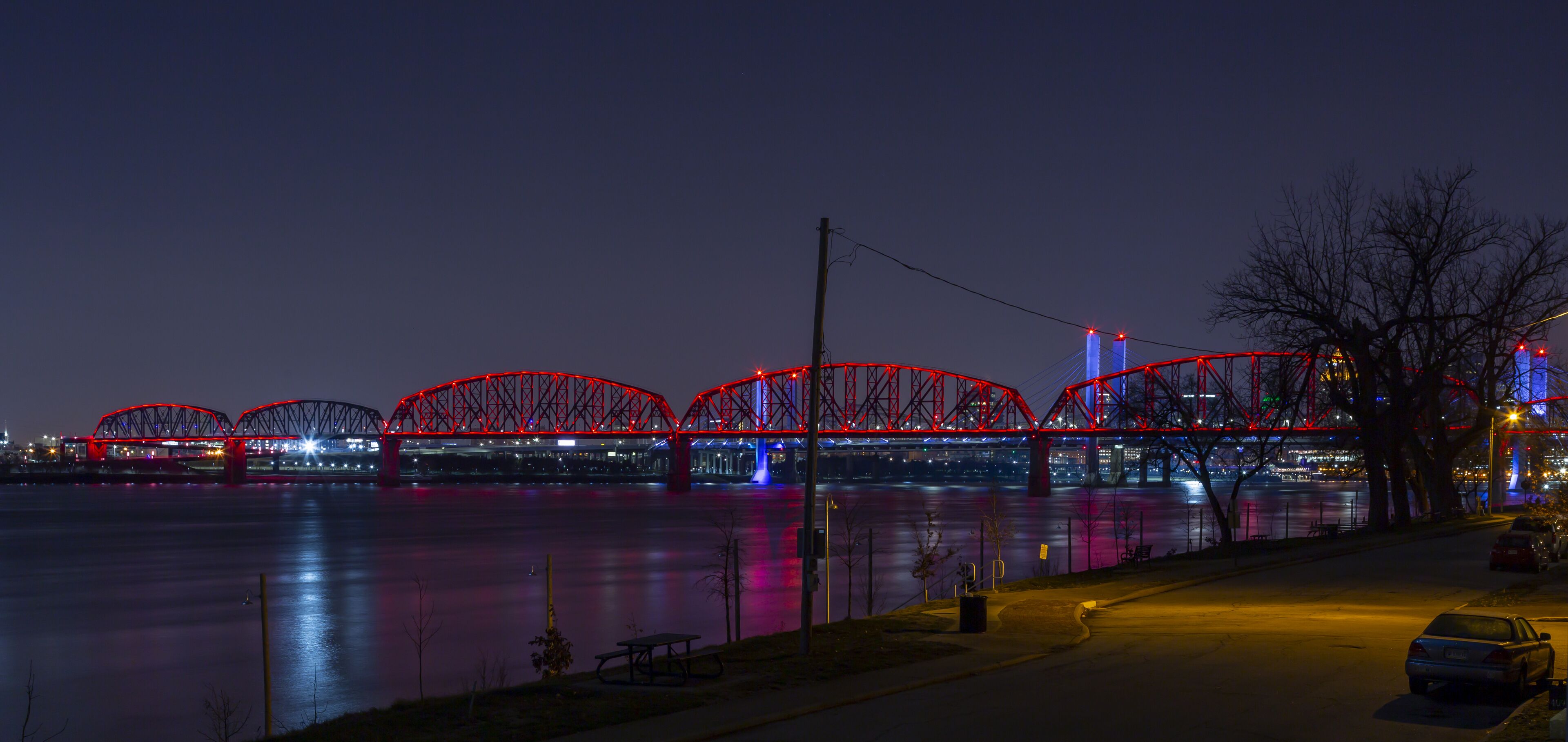 Vista nocturna de un puente iluminado en rojo sobre un río, con una mesa de picnic y bancos a lo largo de la orilla.