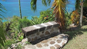 A stone bench overlooking a tropical beach with clear blue water and boats in the distance. - Villa Cap Ouest - Private swimming pool and private sea access - Grande Anse (Les Anses-d'Arlet)