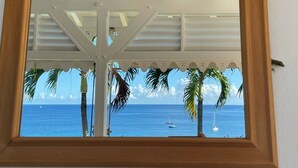 A wooden-framed window with a view of a beach, palm trees, and sailboats on the water. - Villa Cap Ouest - Private swimming pool and private sea access - Grande Anse (Les Anses-d'Arlet)