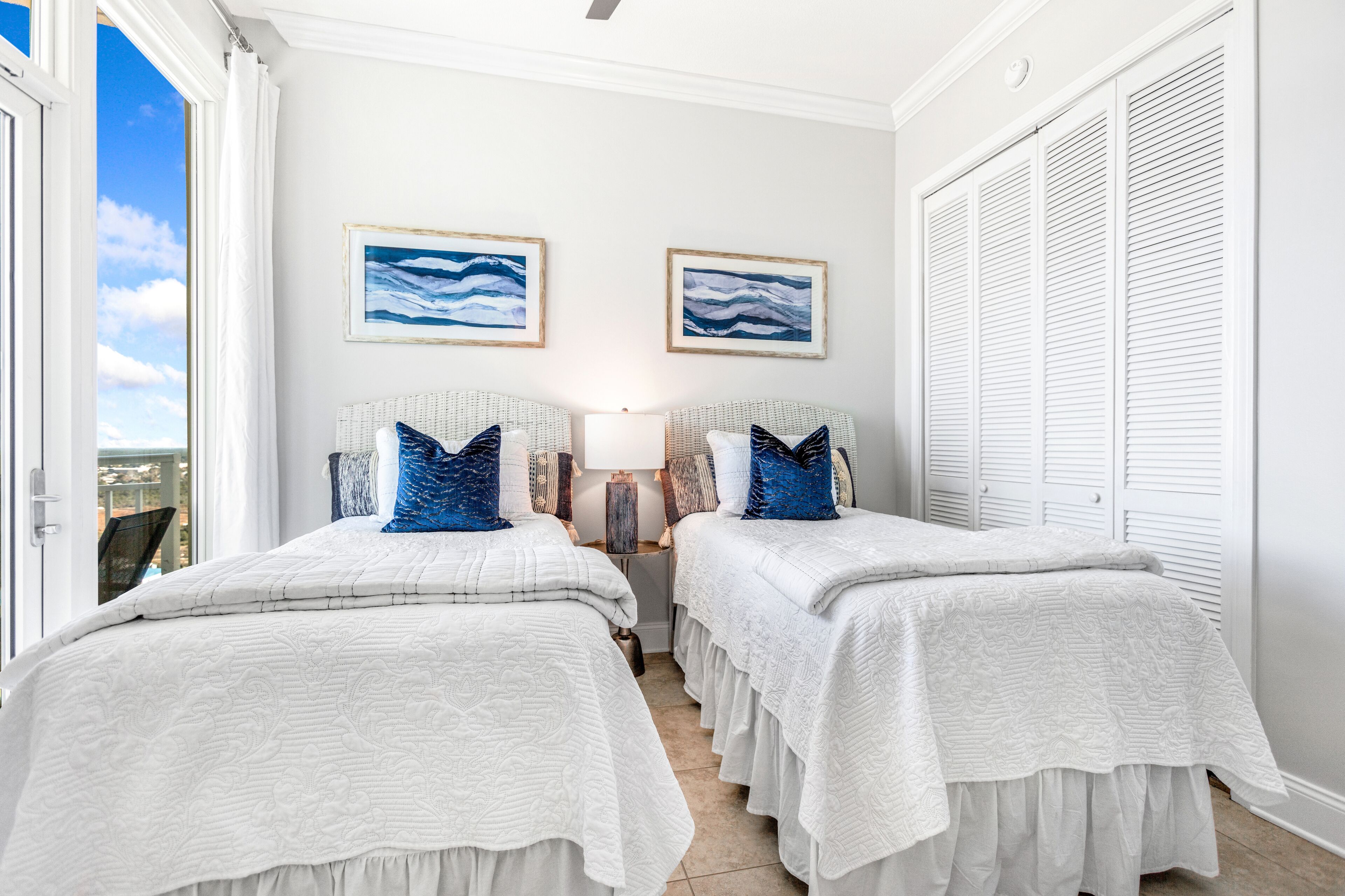 Quarto com duas camas, roupa de cama branca e vista para o oceano através de uma janela grande.