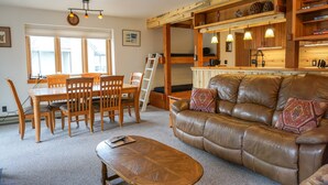 A cozy living room with a brown leather sofa, wooden dining table, and chairs. There is a ladder leaning against the wall and a bookshelf filled with books and decorative items. - JHRL - Aspens Juniper 114, perfect for a couple or single vacationers (Wilson)
