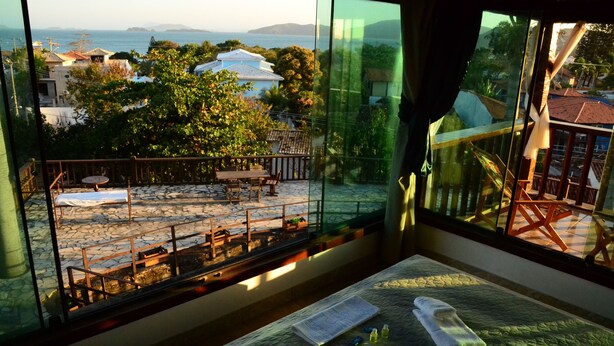Una habitación con un amplio ventanal que da a un patio con mesa y sillas, una cama con colcha y toallas, y vistas al mar y edificios.