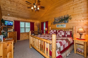 A cozy log cabin bedroom with a wooden bed, a television, a desk, and a sign for 'Band and Tackle'.