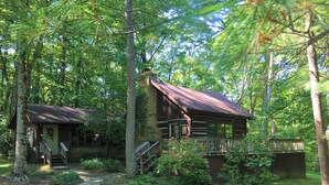 Un chalet rustique doté d’une cheminée en pierre, d’une terrasse en bois et d’un chemin de gravier menant à l’entrée.