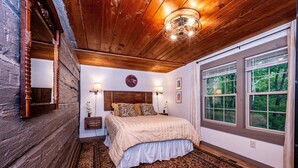 A bedroom with a wooden ceiling, a large bed with a beige bedspread, two wall-mounted lamps, and a large window with curtains. - Walk to Nashville | Hot Tub, Creek Views & Cozy Log Cabin 2BR , 2BA (Nashville)