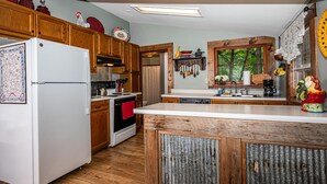 A kitchen with wooden cabinets, a white refrigerator, a wooden island, and a window with a view of trees. - Walk to Nashville | Hot Tub, Creek Views & Cozy Log Cabin 2BR , 2BA (Nashville)