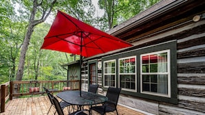 A wooden deck with a table and chairs, a red umbrella, and a cabin with windows and a door. - Walk to Nashville | Hot Tub, Creek Views & Cozy Log Cabin 2BR , 2BA (Nashville)