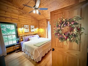 A wooden cabin bedroom with a bed, ceiling fan, wall-mounted lamps, and a window with a view.