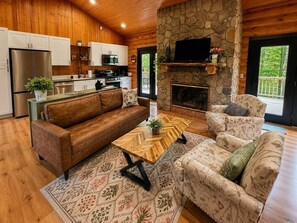 A cozy cabin interior with a wooden ceiling, stone fireplace, a kitchen with modern appliances, a sofa, and a patterned rug.