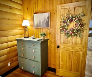 Interior de una cabaña de madera con un aparador, una lámpara, un cuadro enmarcado y una guirnalda en la puerta.