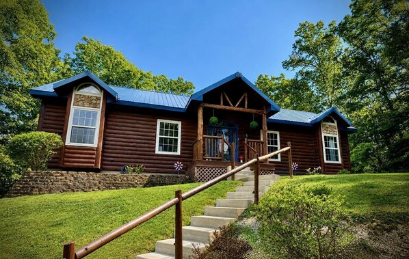 A two-story log cabin with a stone foundation, a wooden porch, and a staircase leading to the entrance.