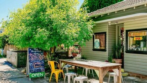Ein kleiner Außenbereich mit Holztisch und -stühlen, ein Kreidetafel mit Werbung für Kräutertees und Bio-Kaffee, und eine Schaufensterfront mit einem Schild, auf dem 'OPEN' steht.