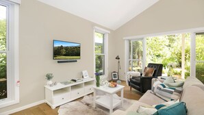 Ein Wohnzimmer mit Wandfernseher, einer weißen Unterhaltungsecke, einem Couchtisch, einer Couch und großen Fenstern mit Blick auf Grünflächen.