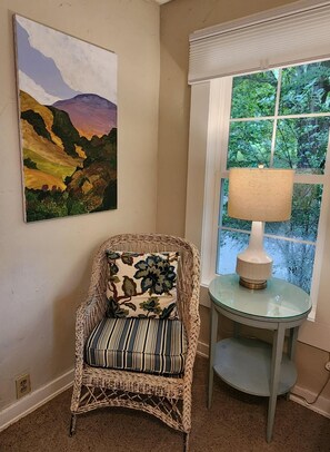 Uma cadeira de vime com um travesseiro estampado e uma almofada listrada, uma mesa de vidro com uma luminária, e uma pintura de paisagem na parede.