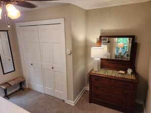 Quarto com porta branca fechada, cômoda de madeira com espelho, um banco e ventilador de teto.