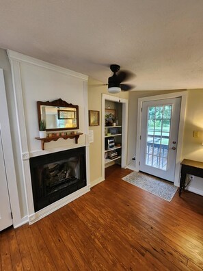 A room with a fireplace, a ceiling fan, a door leading outside, and a bookshelf.