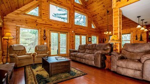 A spacious wooden cabin with large windows, a cozy living room with a sofa and armchairs, and a fireplace. - Private Log Cabin Retreat | Pond, Hot Tub, Game Room, Sleeps 8 (Columbus)