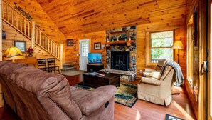 A log cabin interior with a stone fireplace, wooden staircase, and a cozy seating area. - Private Log Cabin Retreat | Pond, Hot Tub, Game Room, Sleeps 8 (Columbus)