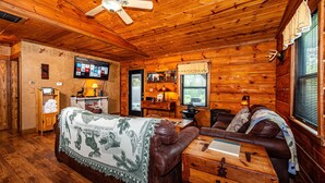 A cozy cabin interior with a wooden ceiling, a ceiling fan, a television mounted on the wall, a fireplace, a sofa, and a wooden dresser. - Cozy Pondside Cabin w/ Hot Tub | Near Bloomington & Nashville (Nashville)