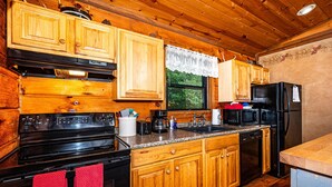 A kitchen with wooden cabinets, a black stove, a refrigerator, and a window with a view of greenery. - Cozy Pondside Cabin w/ Hot Tub | Near Bloomington & Nashville (Nashville)