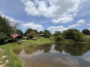 A serene lakeside setting with a wooden gazebo, a small building, and lush greenery. - Cozy Pondside Cabin w/ Hot Tub | Near Bloomington & Nashville (Nashville)