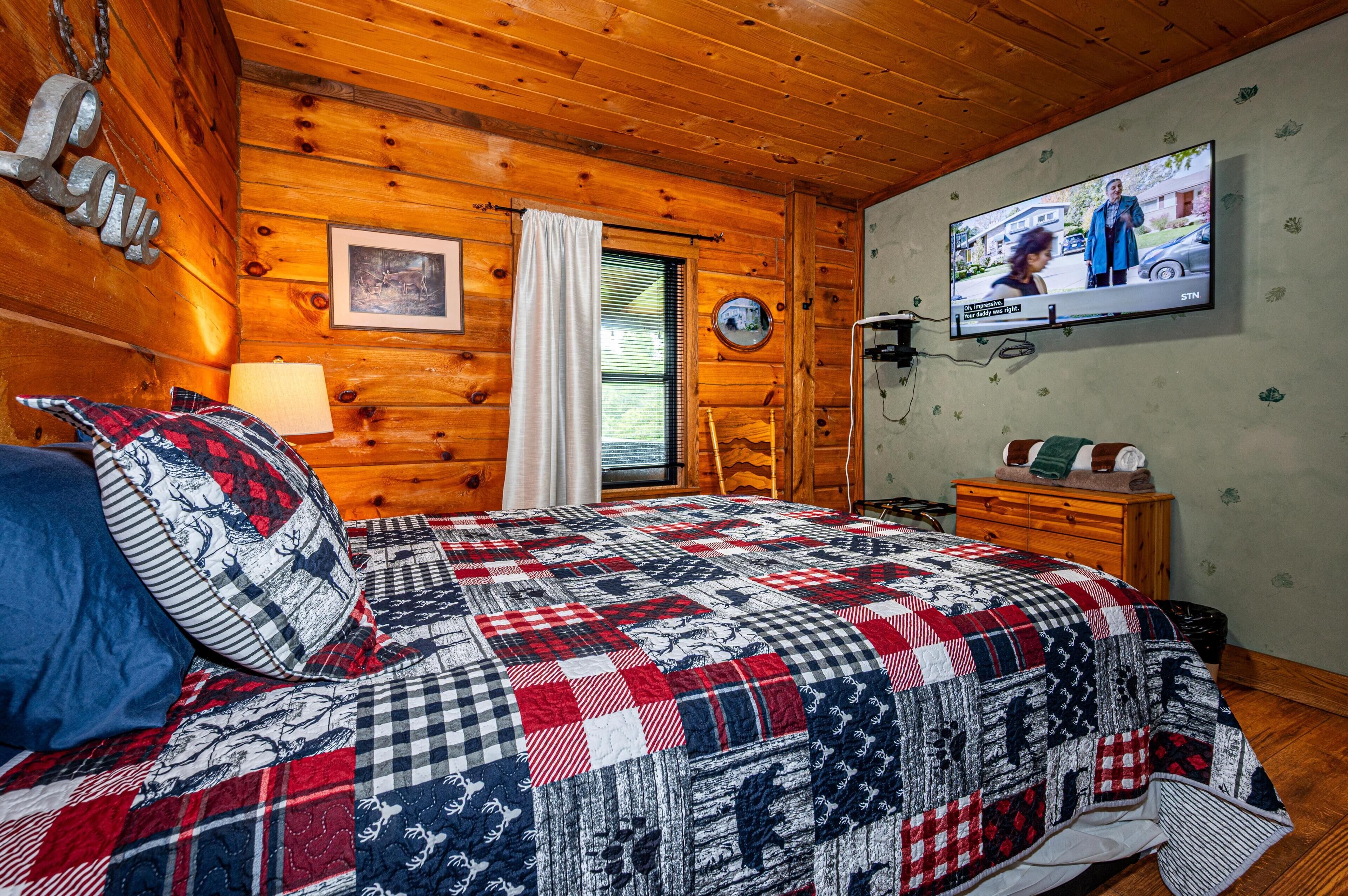 A cozy cabin room with a wooden interior, a bed with a patterned quilt, a television on the wall, and a window with blinds.