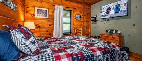 A cozy cabin room with a wooden interior, a bed with a patterned quilt, a television on the wall, and a window with blinds.