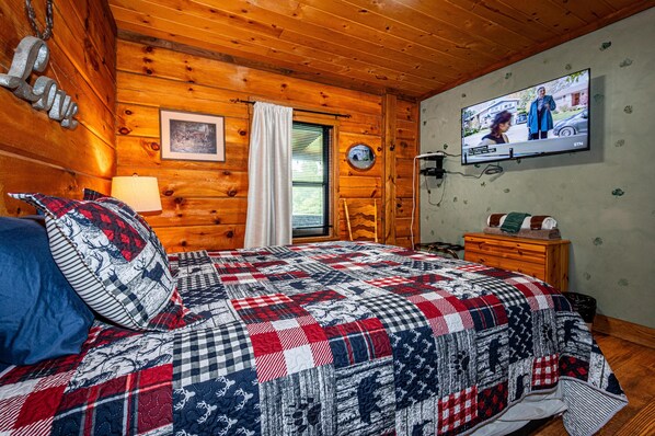 A cozy cabin room with a wooden interior, a bed with a patterned quilt, a television on the wall, and a window with blinds. - Cozy Pondside Cabin w/ Hot Tub | Near Bloomington & Nashville (Nashville)