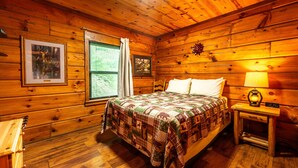 A wooden cabin bedroom with a bed, a nightstand, a lamp, and two framed pictures on the wall. - Cozy Pondside Cabin w/ Hot Tub | Near Bloomington & Nashville (Nashville)